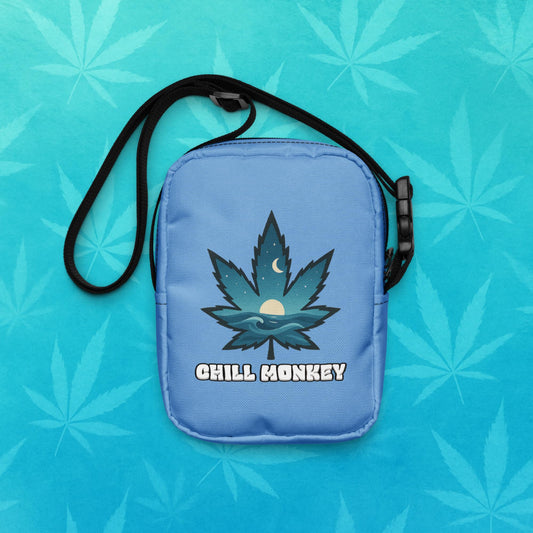 Small blue cross-body utility bag with black adjustable strap, featuring a stylized cannabis leaf filled with ocean waves, moon and sun graphic above the text “Chill Monkey”; back view on aqua leaf backdrop.