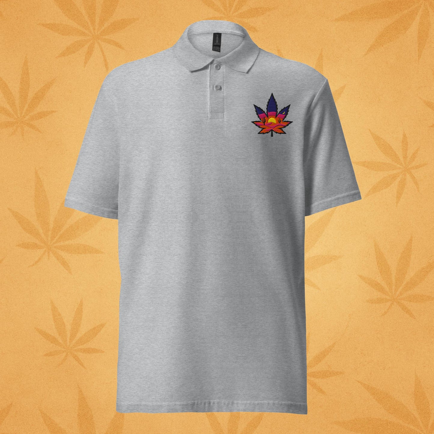 Sport-grey pique polo shirt with a small embroidered sunset-colored cannabis leaf emblem on the left chest; front view against a warm desert-orange leaf backdrop.