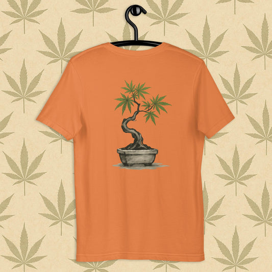 Back view of a orange unisex Bella+Canvas tee showcasing a large hand-painted cannabis-bonsai illustration in Japanese sumi-e style, centered on the shirt against a neutral leaf backdrop.