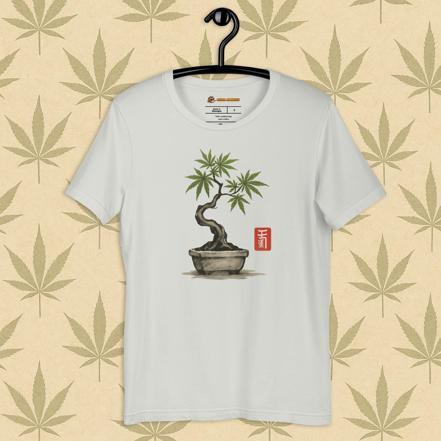 Front view of a silver unisex Bella+Canvas tee featuring a hand-drawn cannabis-bonsai pot with Japanese-style brushwork and a small red hanko seal, shown on a neutral leaf backdrop.