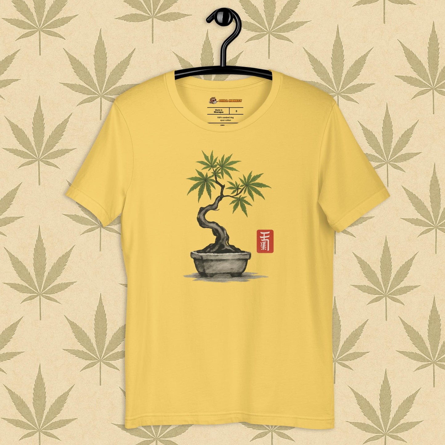 Front view of a yellow unisex Bella+Canvas tee featuring a hand-drawn cannabis-bonsai pot with Japanese-style brushwork and a small red hanko seal, shown on a neutral leaf backdrop.