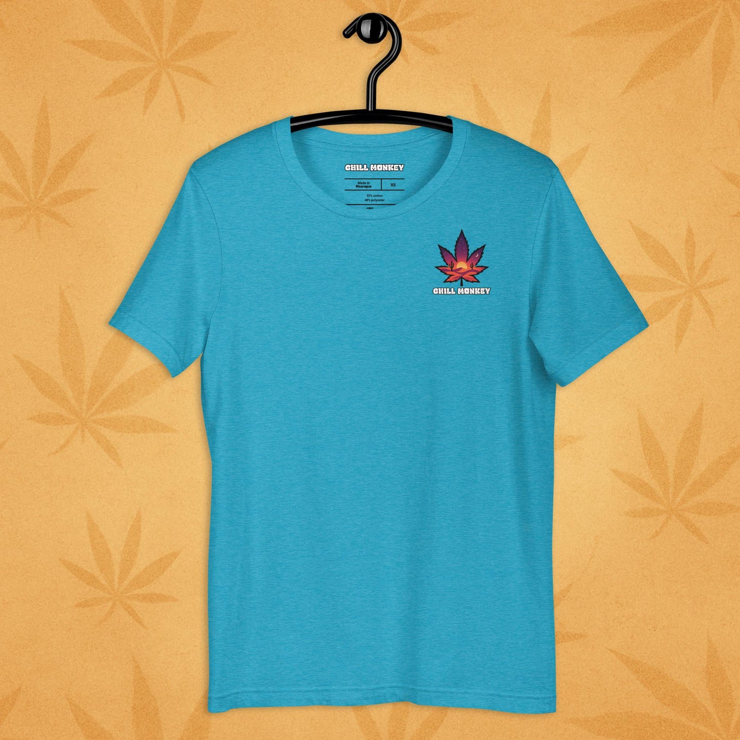 Heather-blue unisex T-shirt with a small, multicolored desert-sunset cannabis leaf emblem on the left chest; front view on a sandy, boho-leaf backdrop.