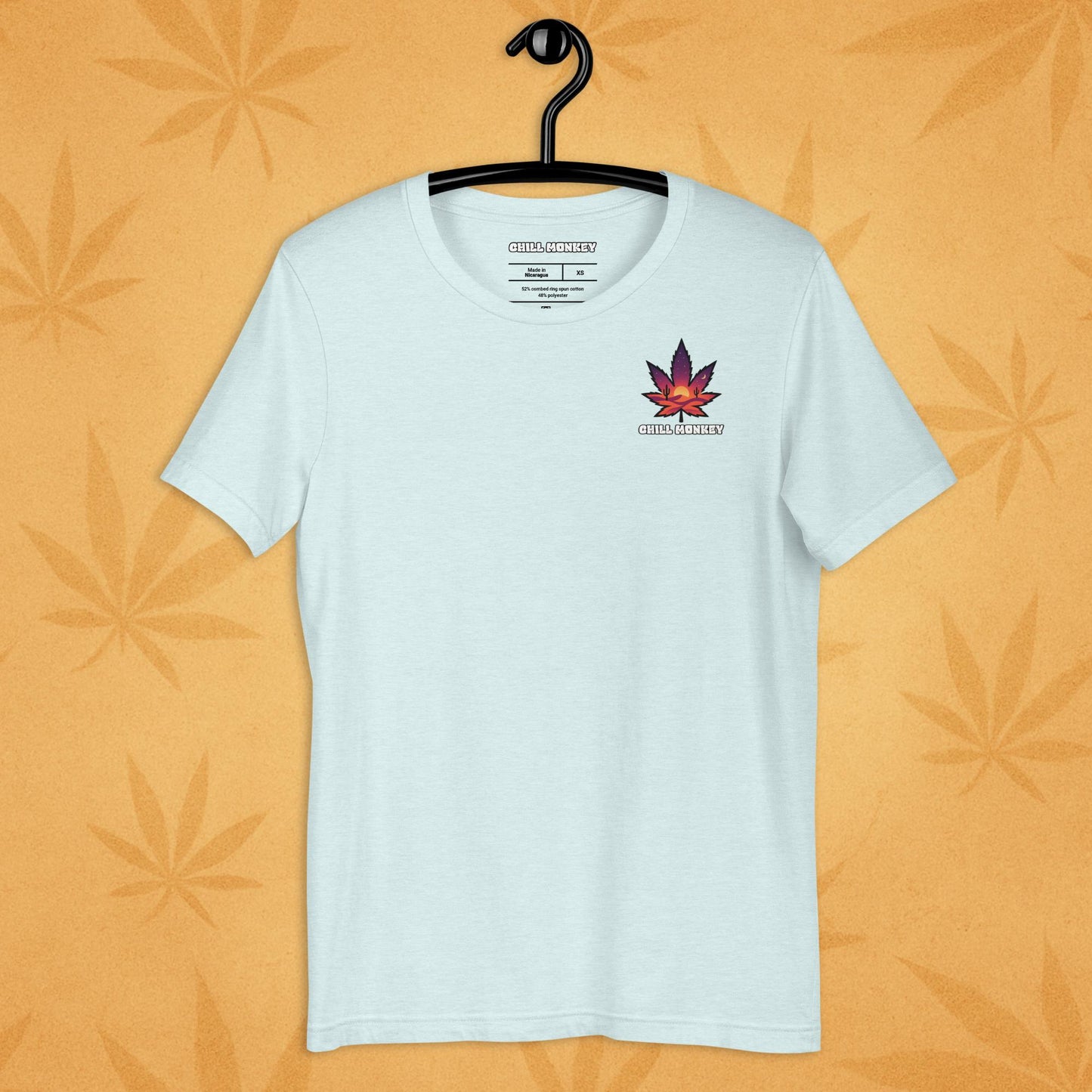 Heather-ice unisex T-shirt with a small, multicolored desert-sunset cannabis leaf emblem on the left chest; front view on a sandy, boho-leaf backdrop.