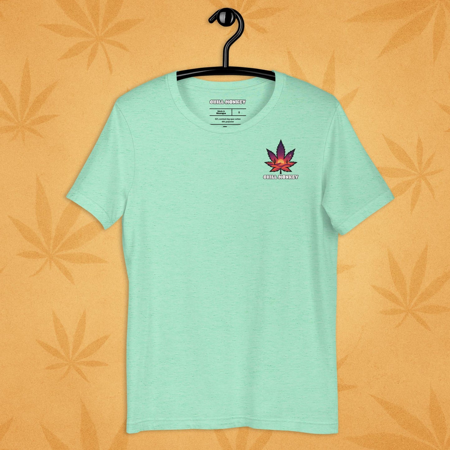 Heather-mint unisex T-shirt with a small, multicolored desert-sunset cannabis leaf emblem on the left chest; front view on a sandy, boho-leaf backdrop.