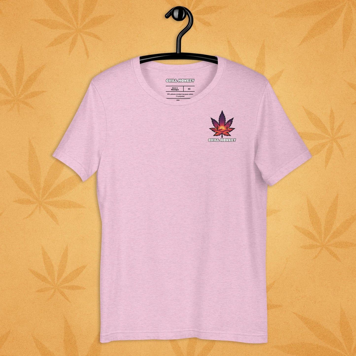Heather-pink unisex T-shirt with a small, multicolored desert-sunset cannabis leaf emblem on the left chest; front view on a sandy, boho-leaf backdrop.