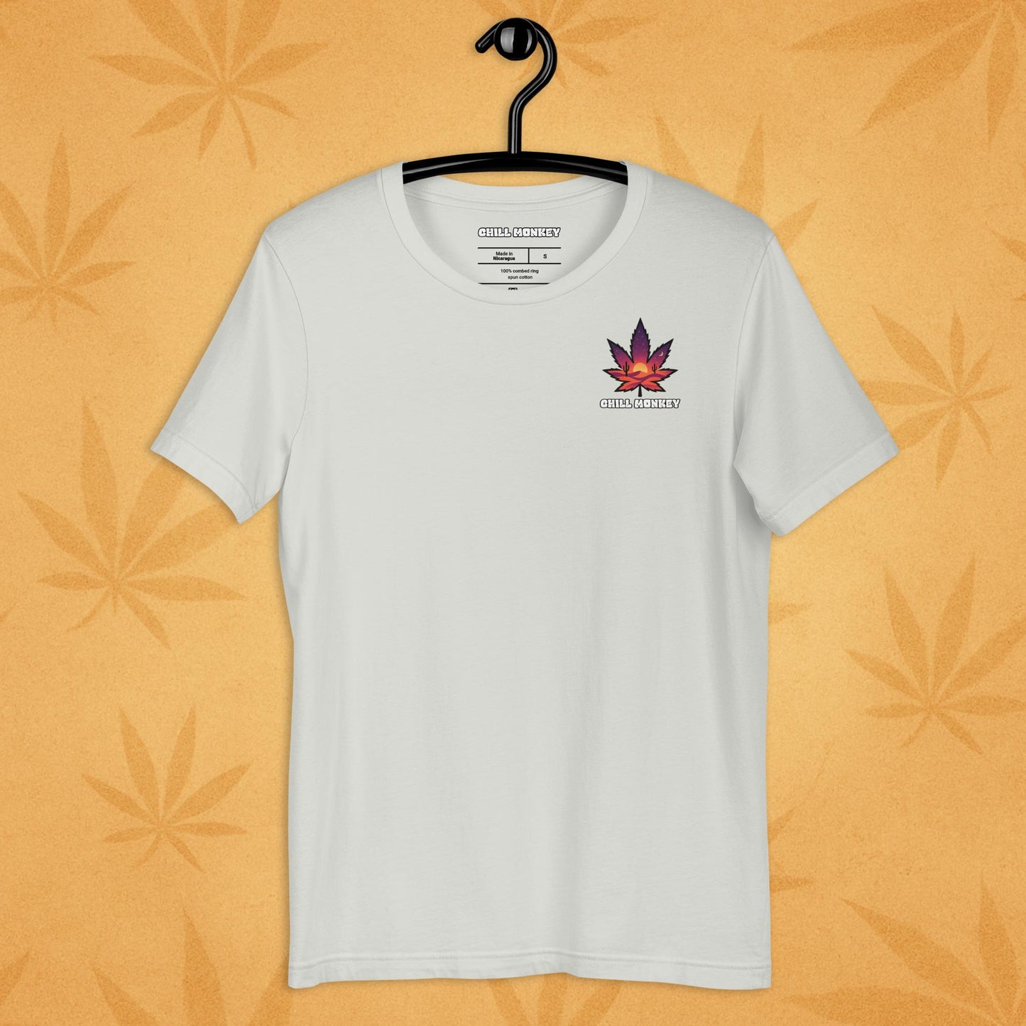 Heather-silver unisex T-shirt with a small, multicolored desert-sunset cannabis leaf emblem on the left chest; front view on a sandy, boho-leaf backdrop.
