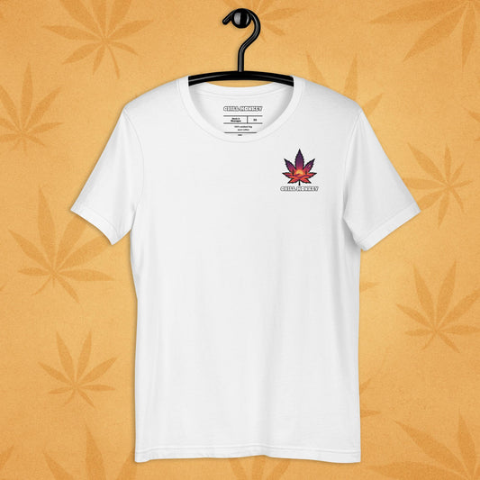 Heather-white unisex T-shirt with a small, multicolored desert-sunset cannabis leaf emblem on the left chest; front view on a sandy, boho-leaf backdrop.