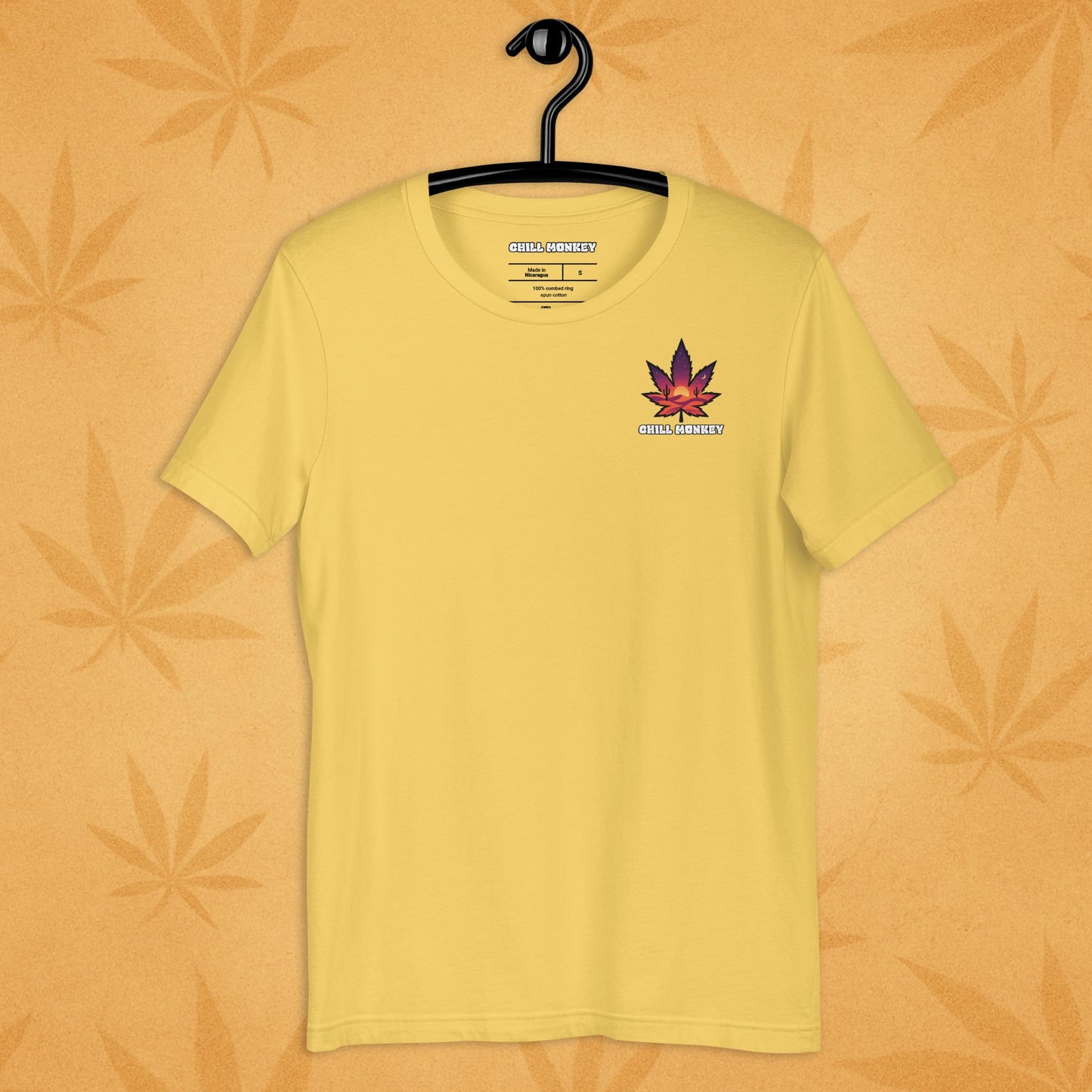 Heather-yellow unisex T-shirt with a small, multicolored desert-sunset cannabis leaf emblem on the left chest; front view on a sandy, boho-leaf backdrop.