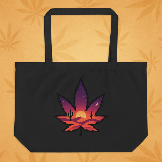 Large oyster-black eco tote bag with a bold, purple-to-orange desert-sunset cannabis leaf graphic on the front, shown against a sandy boho backdrop (front view).