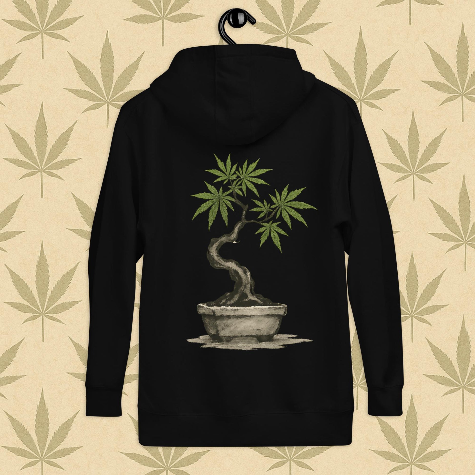 Back view of a black-colored unisex premium hoodie featuring a large Japanese sumi-e style cannabis-bonsai illustration centered on the garment against a neutral leaf backdrop.