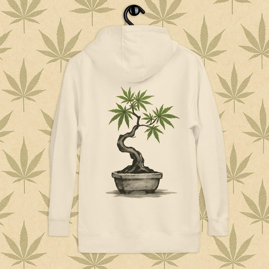 Back view of a bone-colored unisex premium hoodie featuring a large Japanese sumi-e style cannabis-bonsai illustration centered on the garment against a neutral leaf backdrop.
