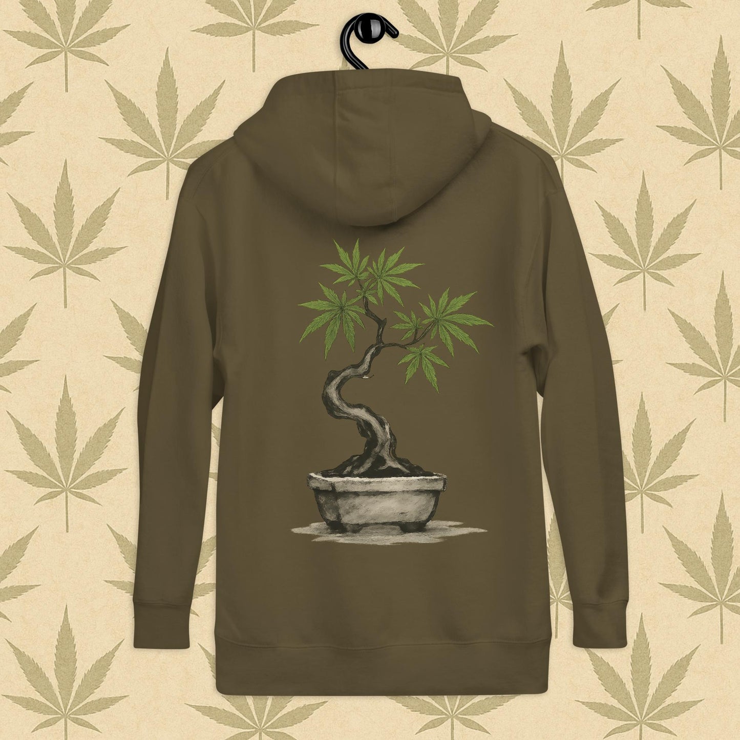 Back view of a green-colored unisex premium hoodie featuring a large Japanese sumi-e style cannabis-bonsai illustration centered on the garment against a neutral leaf backdrop.