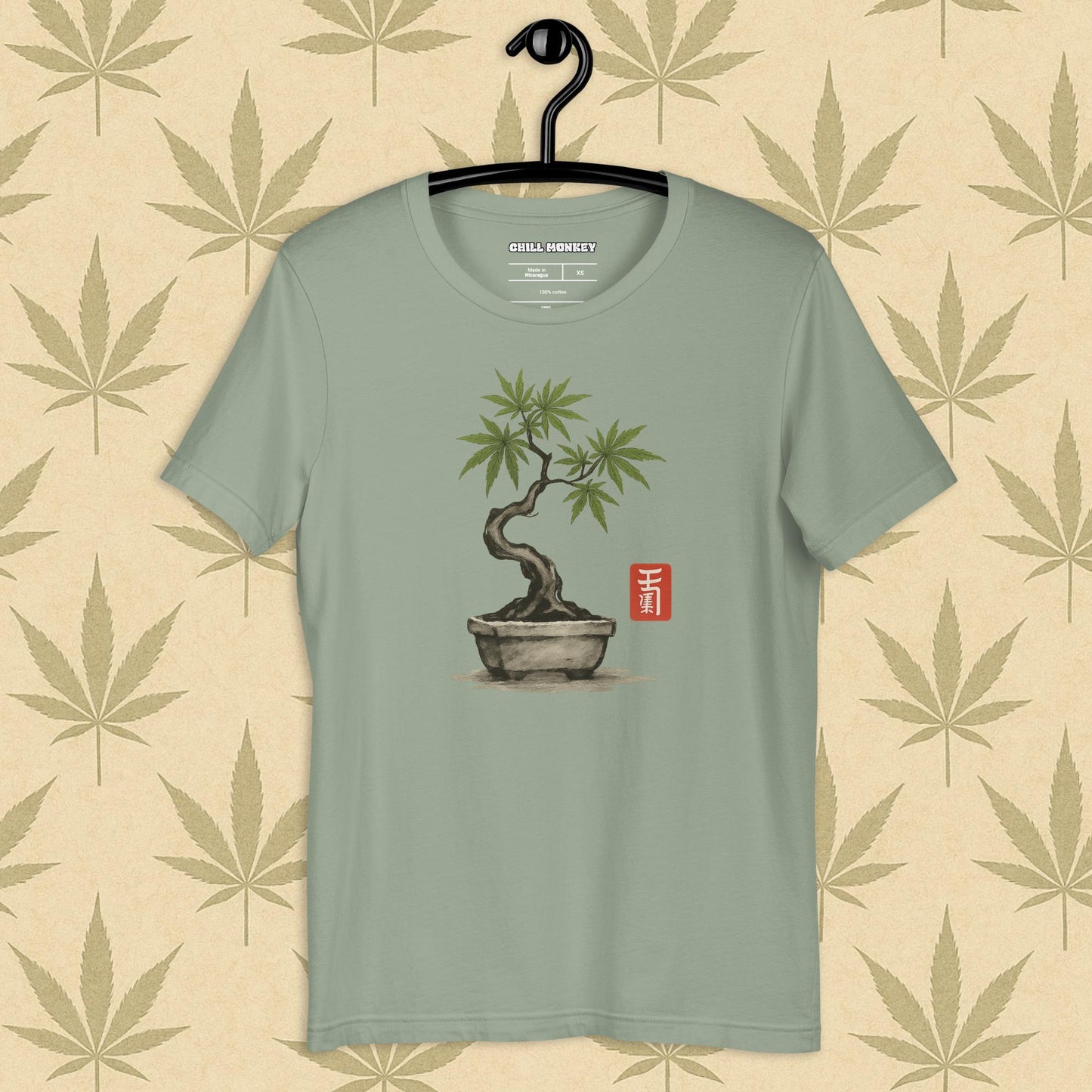 Front view of a grey unisex Bella+Canvas tee featuring a hand-drawn cannabis-bonsai pot with Japanese-style brushwork and a small red hanko seal, shown on a neutral leaf backdrop.