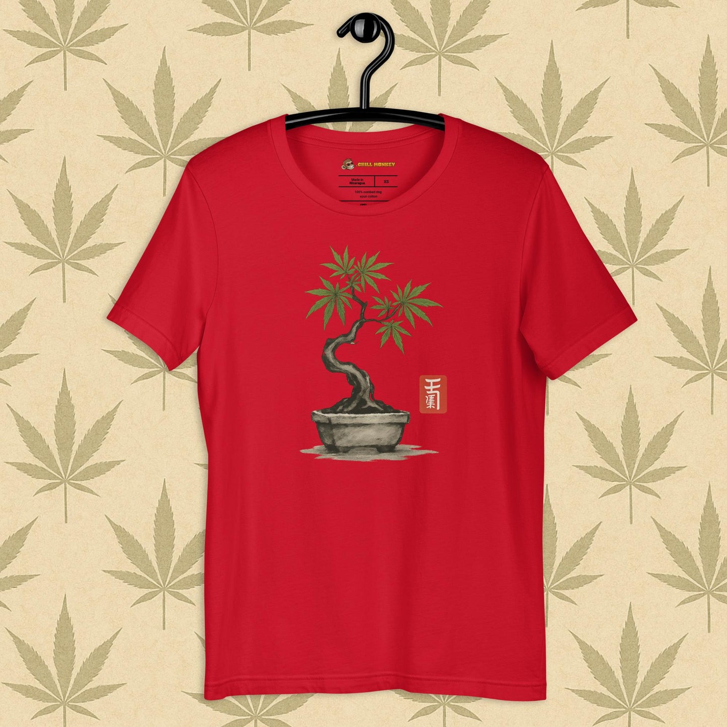 Front view of a red unisex Bella+Canvas tee featuring a hand-drawn cannabis-bonsai pot with Japanese-style brushwork and a small red hanko seal, shown on a neutral leaf backdrop.