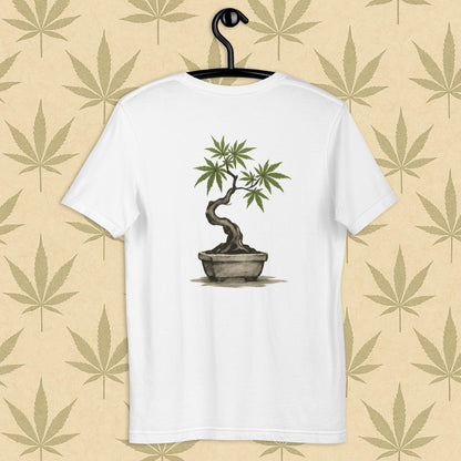 Back view of a white unisex Bella+Canvas tee showcasing a large hand-painted cannabis-bonsai illustration in Japanese sumi-e style, centered on the shirt against a neutral leaf backdrop.