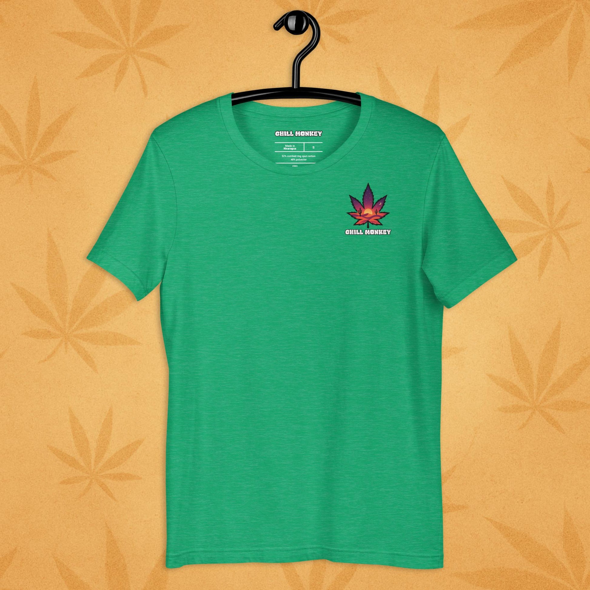 Heather-kelly unisex T-shirt with a small, multicolored desert-sunset cannabis leaf emblem on the left chest; front view on a sandy, boho-leaf backdrop.