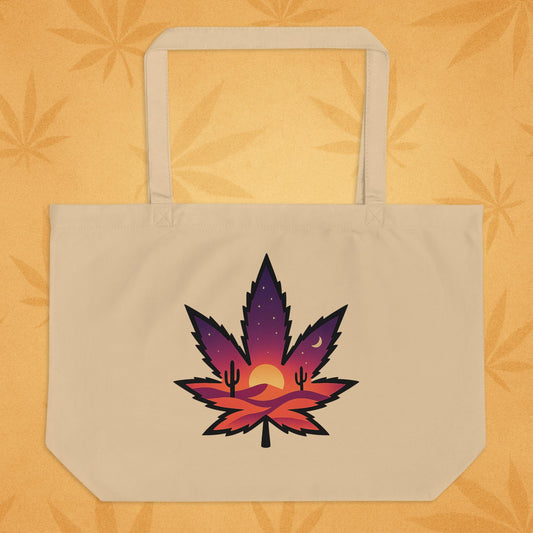 Large oyster-tone eco tote bag with a bold, purple-to-orange desert-sunset cannabis leaf graphic on the front, shown against a sandy boho backdrop (front view).